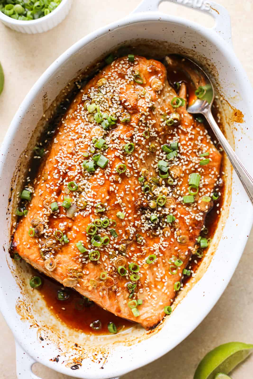 Large salmon filet in white oval baking dish covered in marinade, sesame seeds, and sliced green onions.