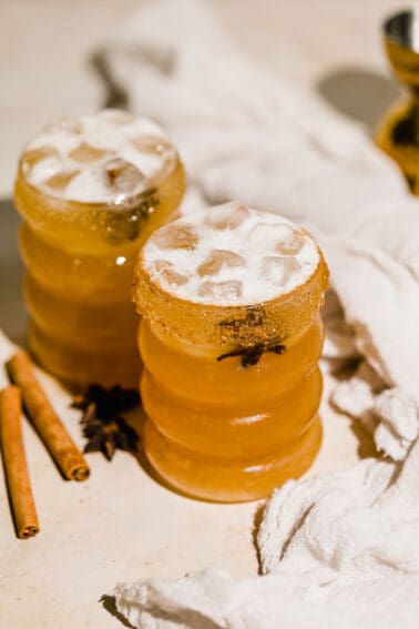 Two Spiced Apple Cider Margarita with cloth napkin laying next to them and cinnamon sticks scattered.