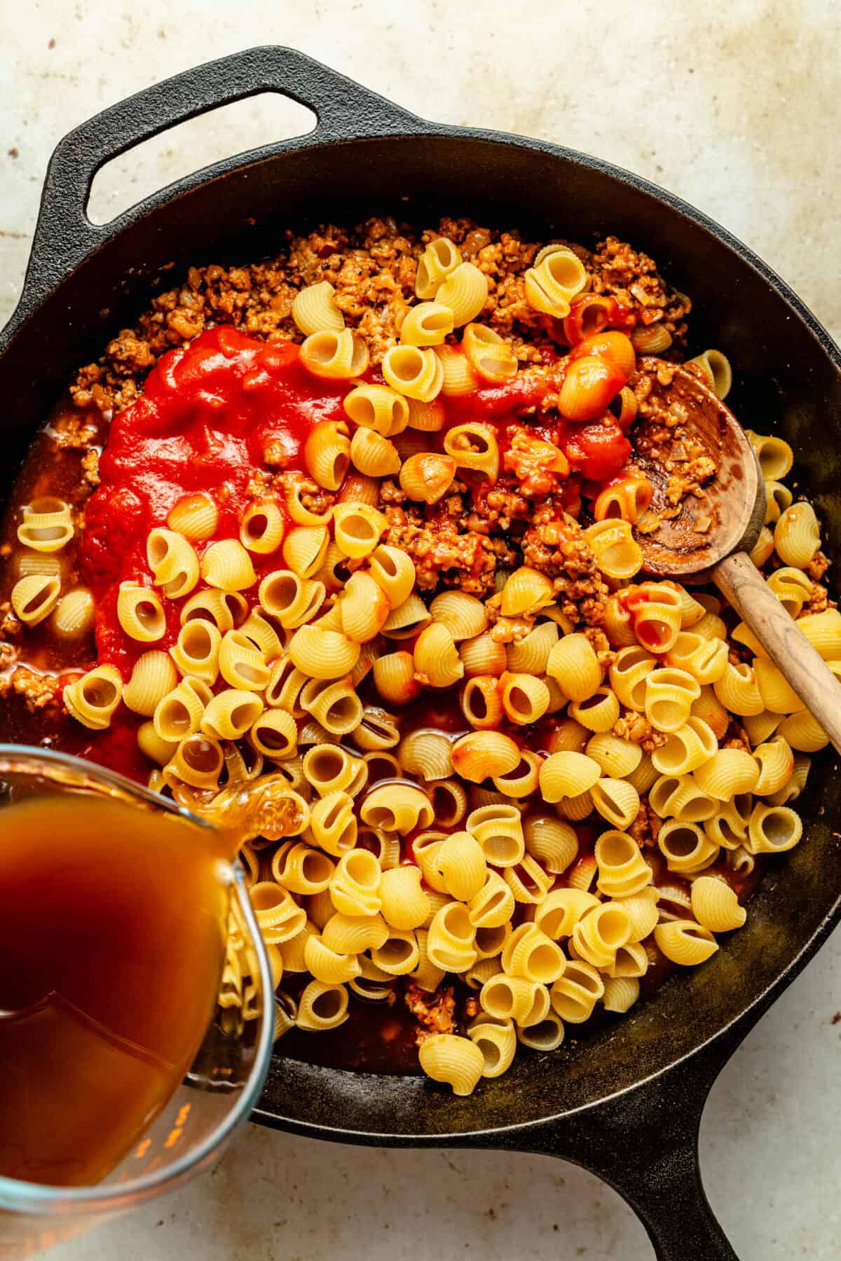 Black cast iron skillet filled with browned ground beef, dry pasta shells, red sauce and broth pouring in.