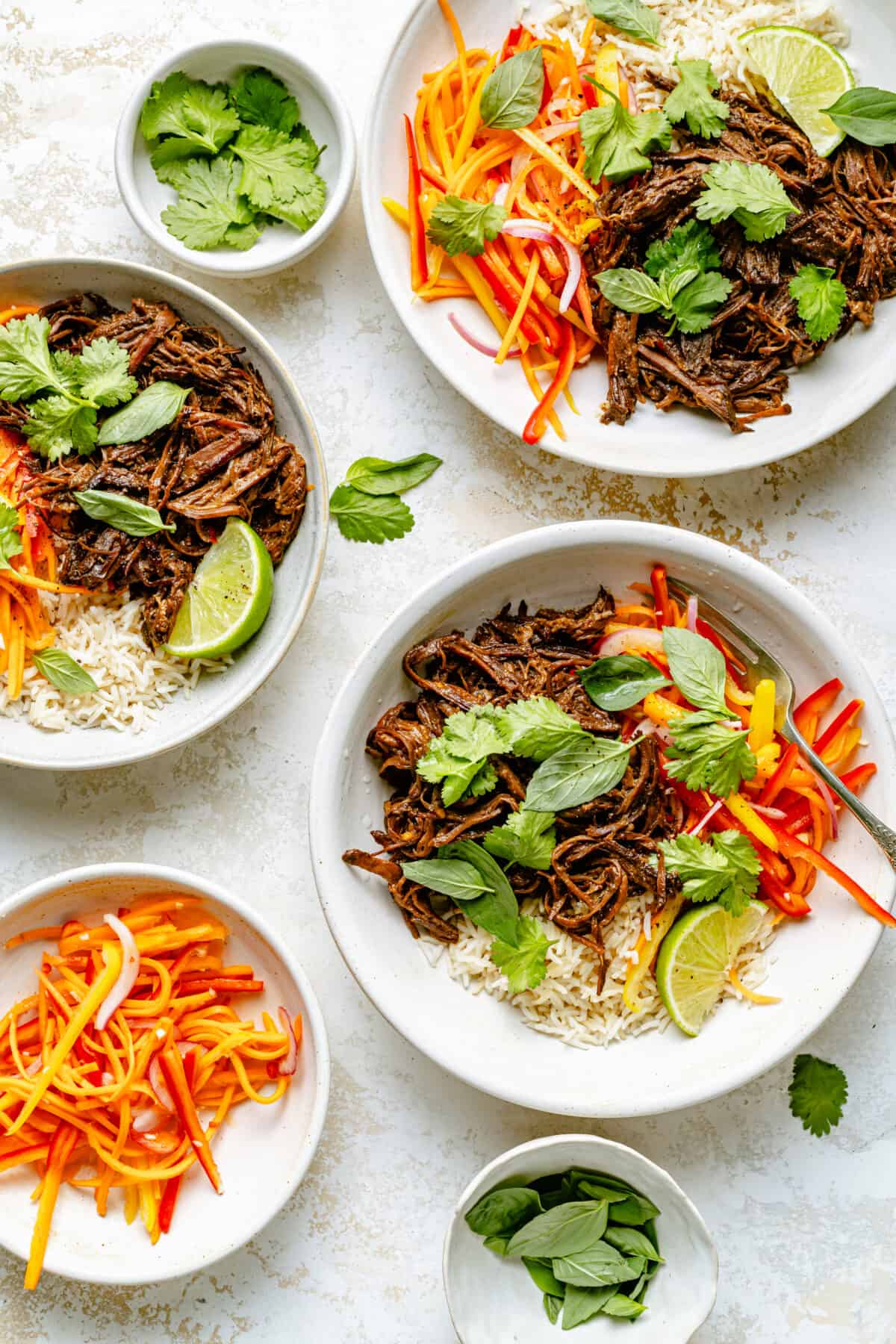 Three bowls with beef, rice, and veggie slaw, topped with herbs on a white stone backdrop.