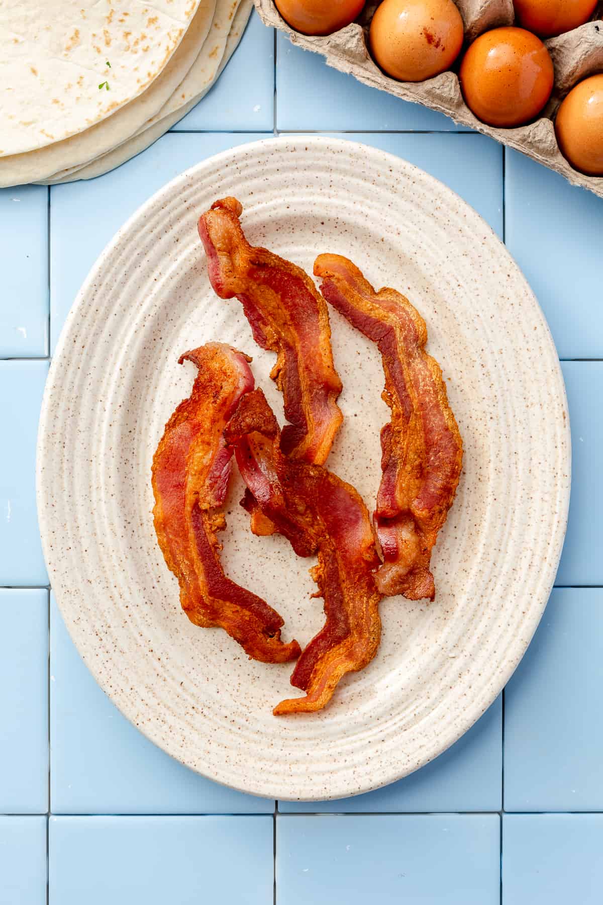 Oval cream plate filled with 4 pieces of crisped bacon on blue tile backdrop.