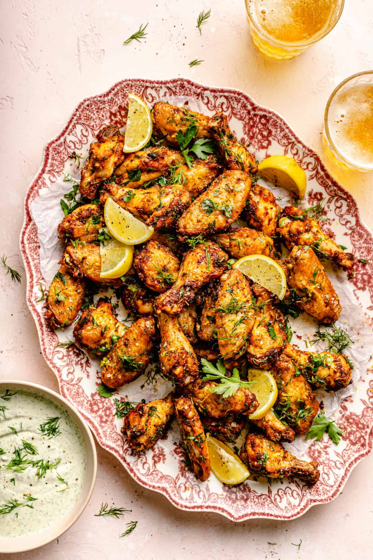 Greek-Inspired Chicken Wings piled on red and white platter with lemon slices on top. Small bowl of feta dipping sauce in bottom corner.