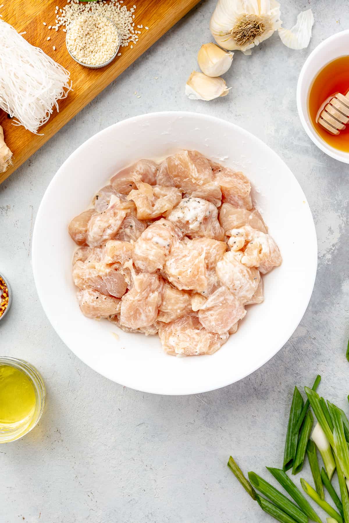 White bowl filled of marinating chicken with other ingredients scattered around it.