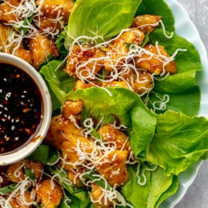 Close up of Four Honey Sesame Chicken Lettuce Cups on a round plate with a small bowl of extra sauce.