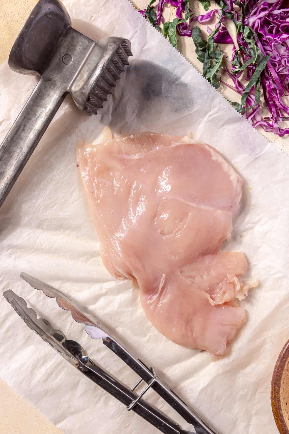 Raw chicken breast on sheet of parchment with meat mallet next to it.