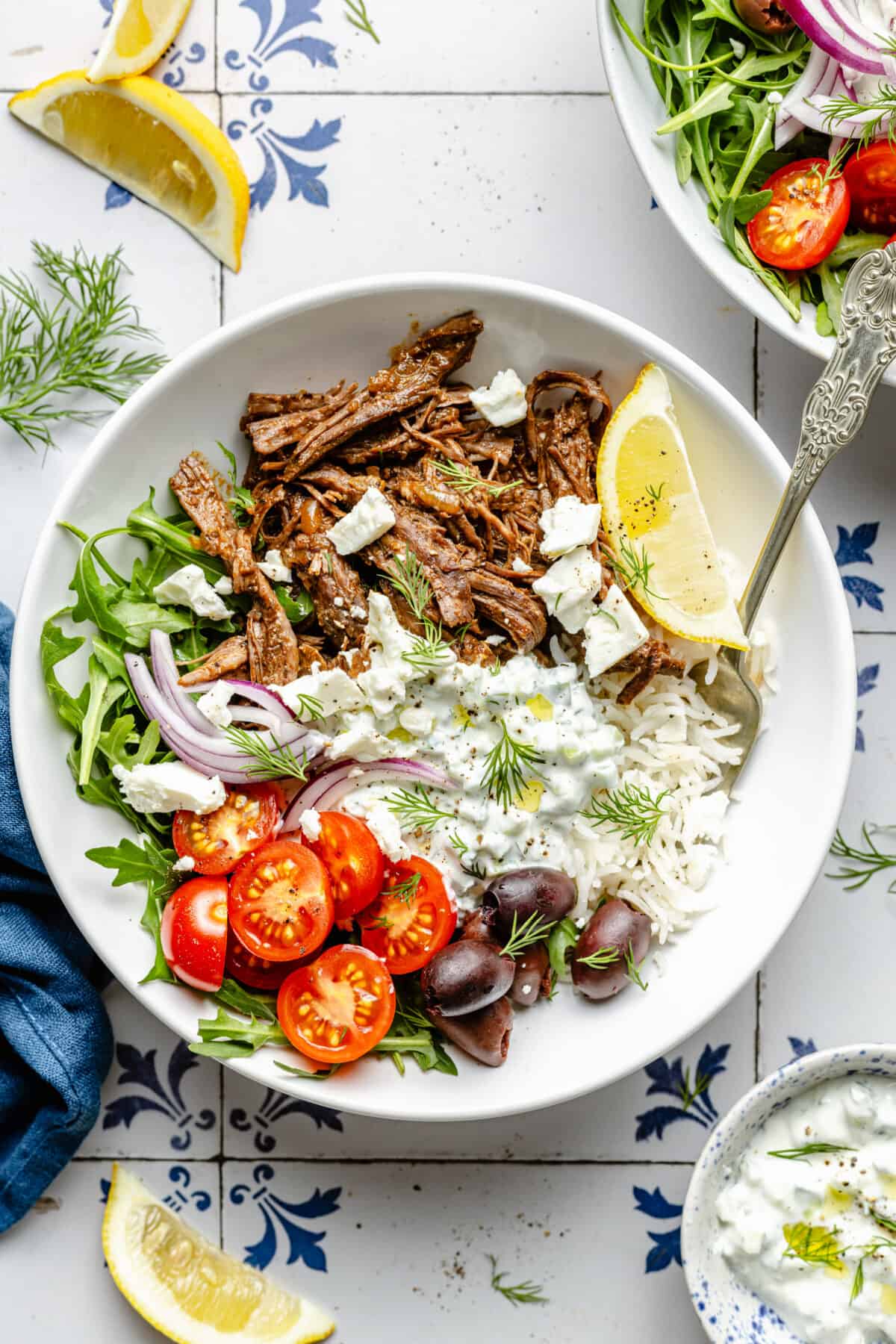 Instant Pot Shredded Greek Beef in bowl with garnishes scattered around.