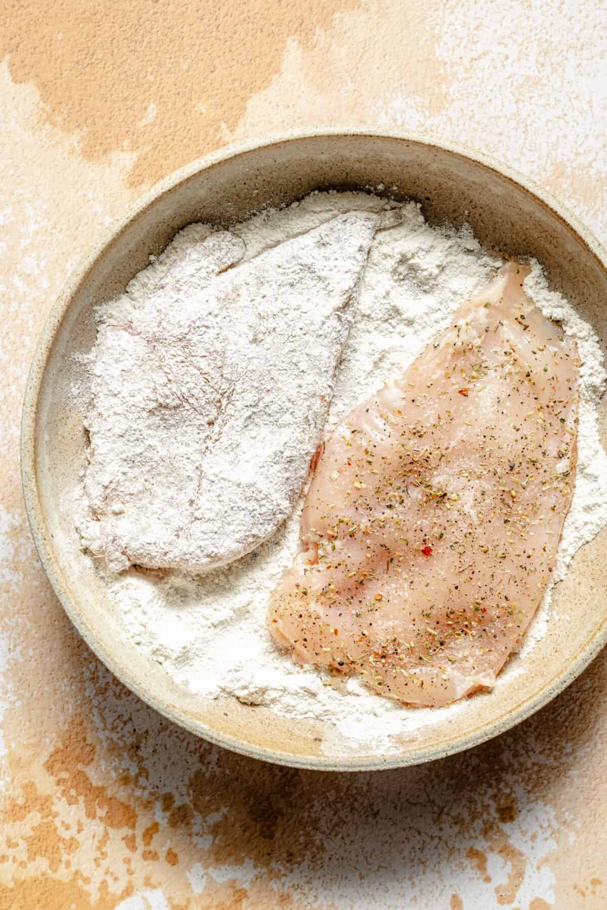 Raw chicken breasts in low bowl filled with flour.