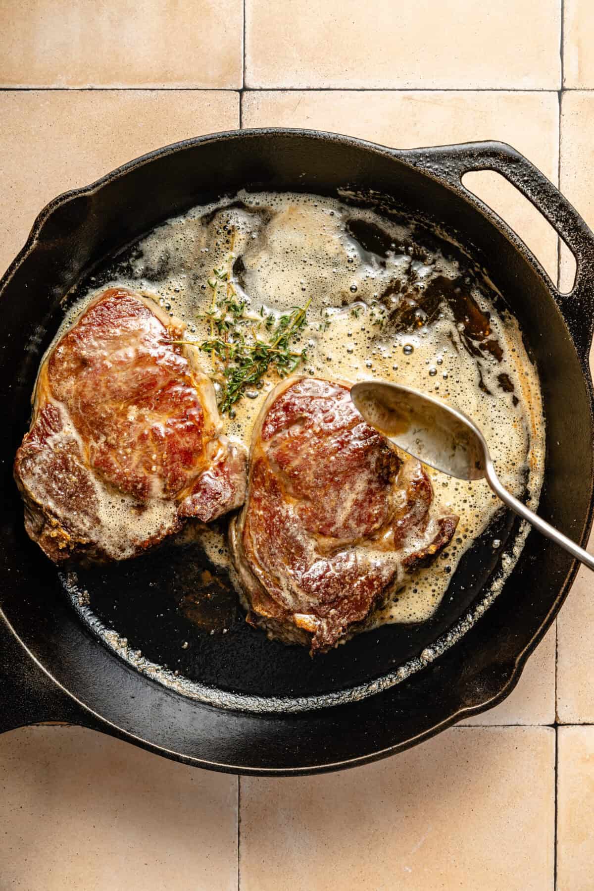 two seared ribeye steaks in a cast iron skillet with melted butter and fresh herbs