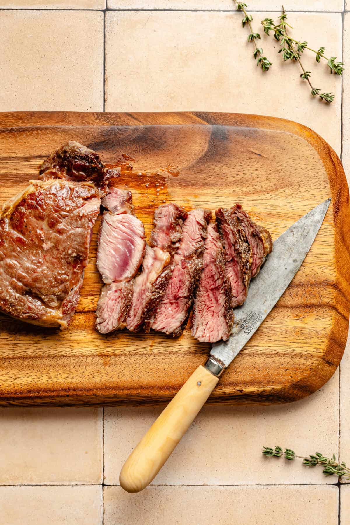 a sliced ribeye steak on a wooden cutting board with fresh herbs and a knife