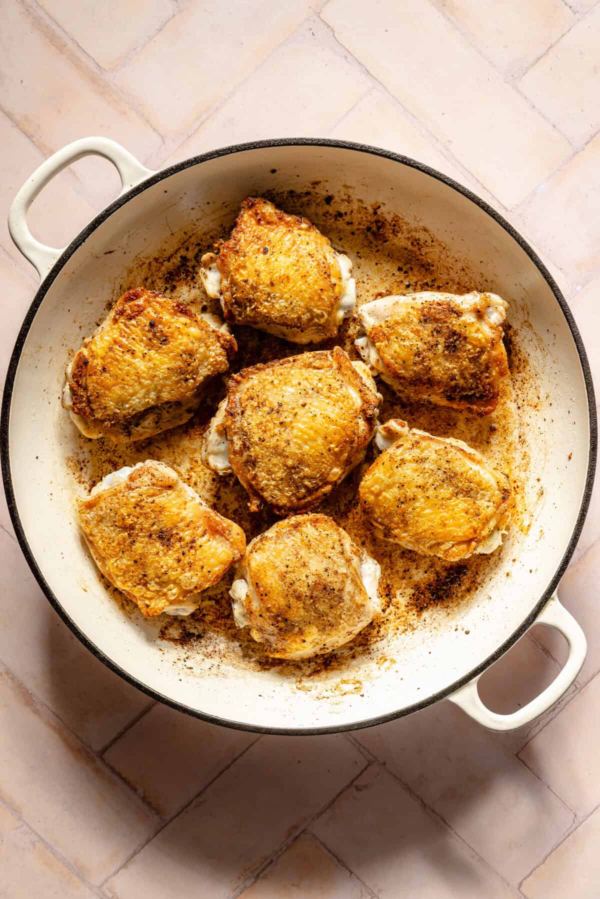 seared chicken thighs in a white Dutch oven on a light pink background