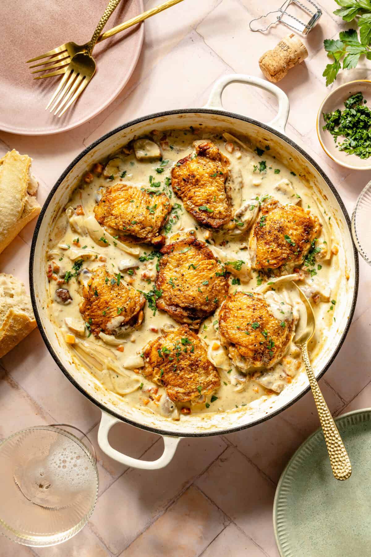 a white Dutch oven with seared chicken in a creamy sauce on a light pink background