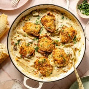a white Dutch oven with seared chicken in a creamy sauce on a light pink background