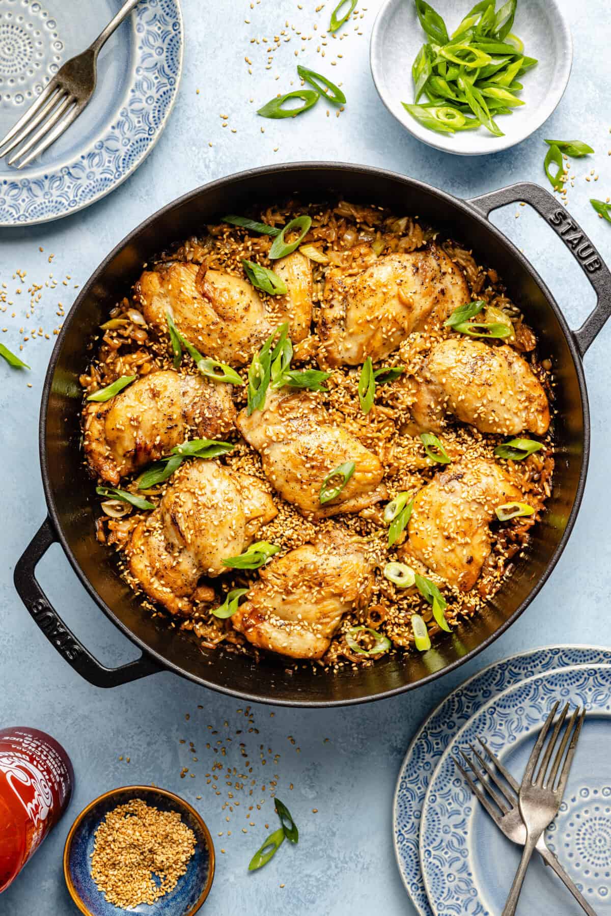 a cast-iron skillet with sesame chicken thighs, rice, and scallions on a light blue background
