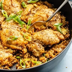a large spoon in a cast-iron skillet with sesame chicken thighs, rice, and sliced green onions