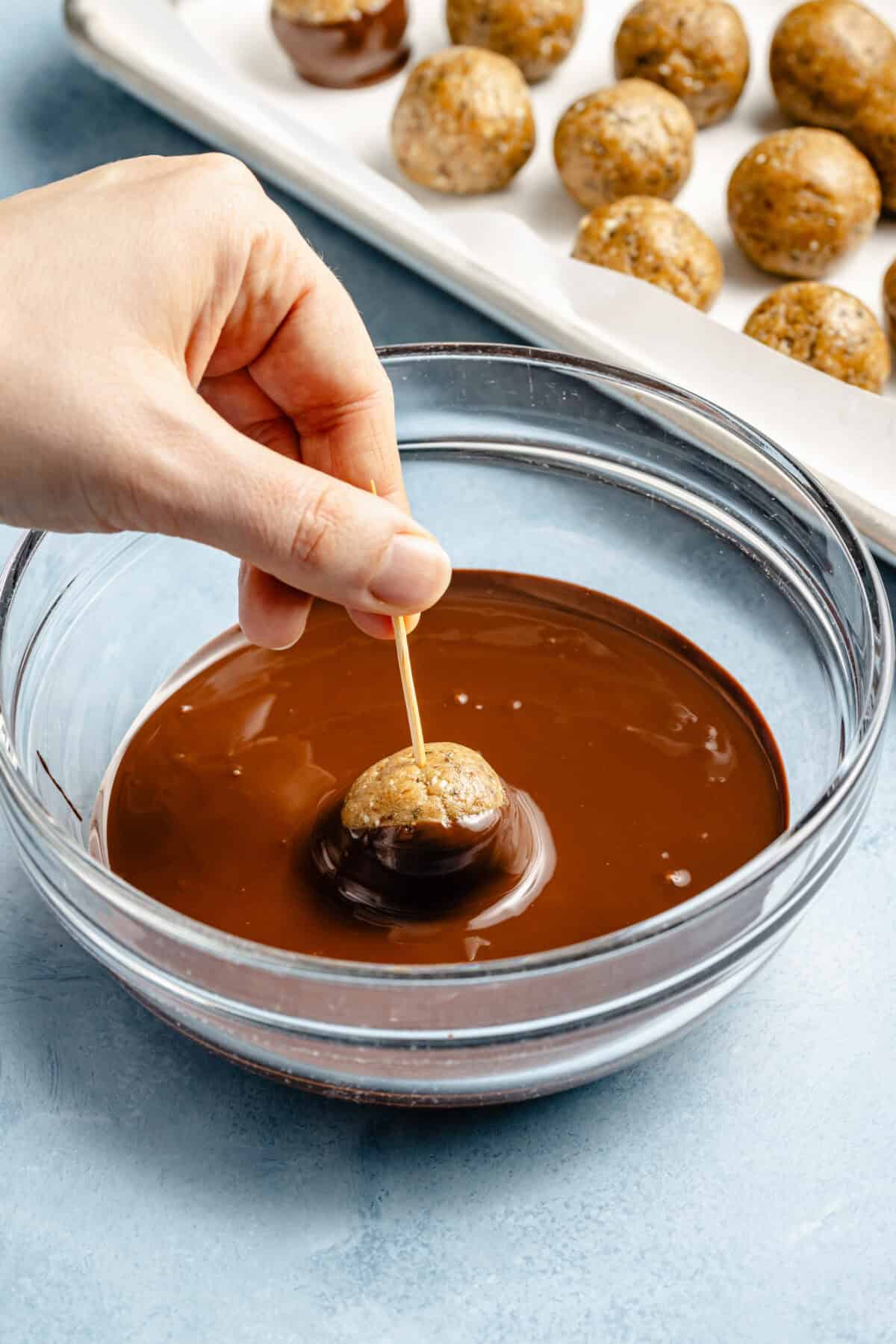 someone dipping a protein ball in melted chocolate in a glass bowl on a light blue background