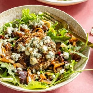 a large white and tan bowl filled with salad greens, caramelized onions, and blue cheese-topped burgers