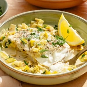 an up-close photo of halibut, leeks, fresh herbs, and lemon wedges in a low bowl