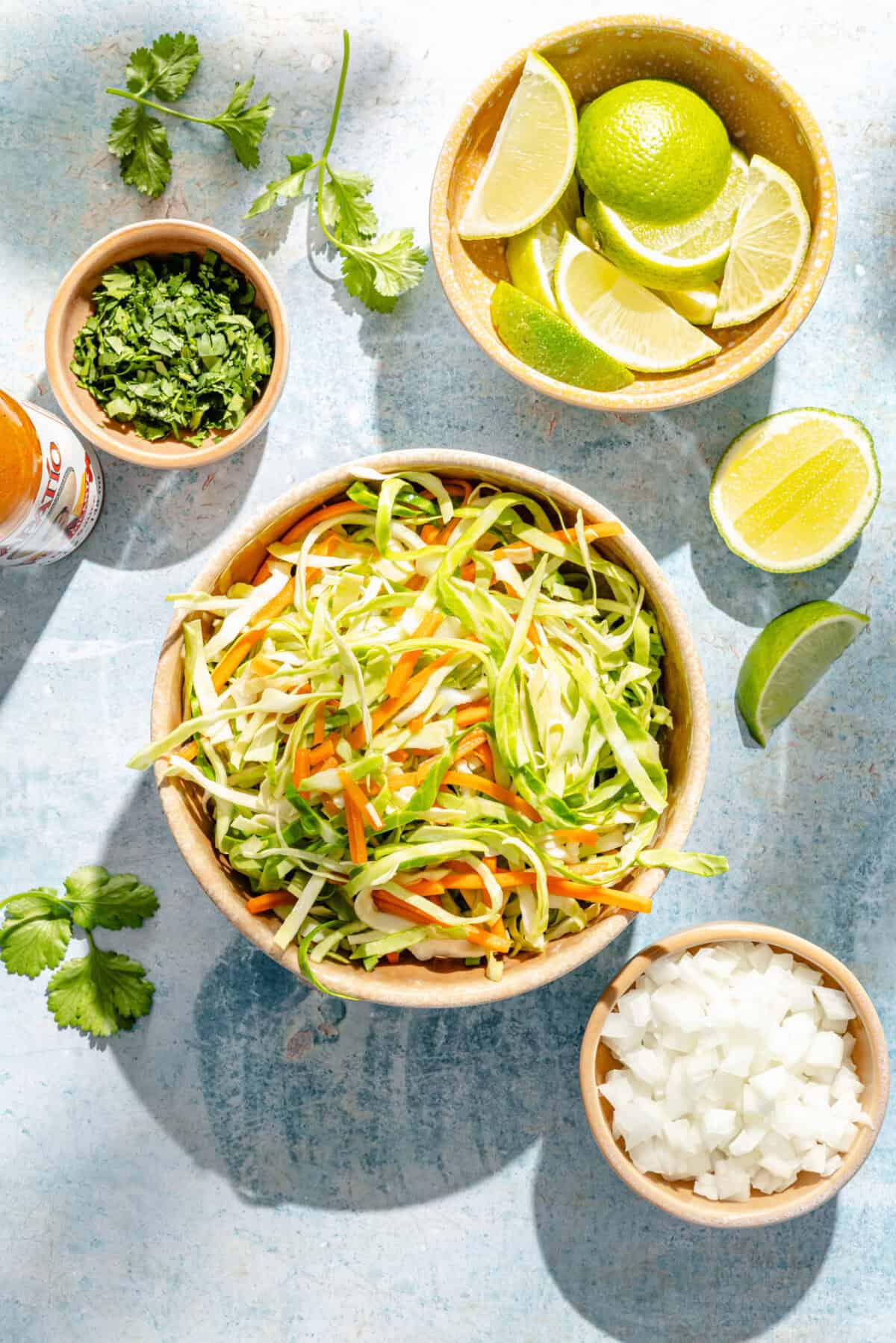 a bowl of cabbage and carrot slaw with limes, white onions, and cilantro on a blue background
