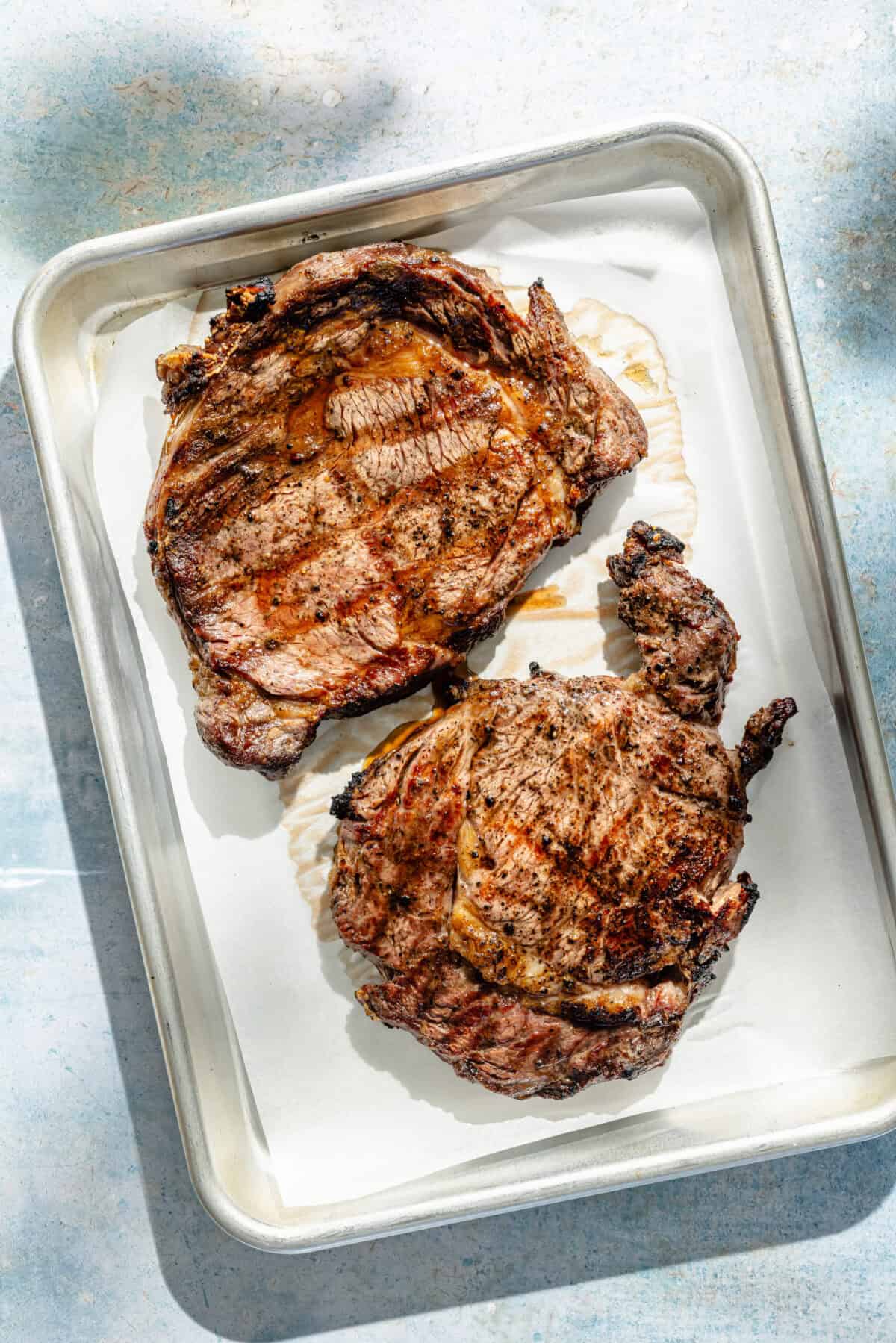 two grilled ribeye steaks on a baking sheet