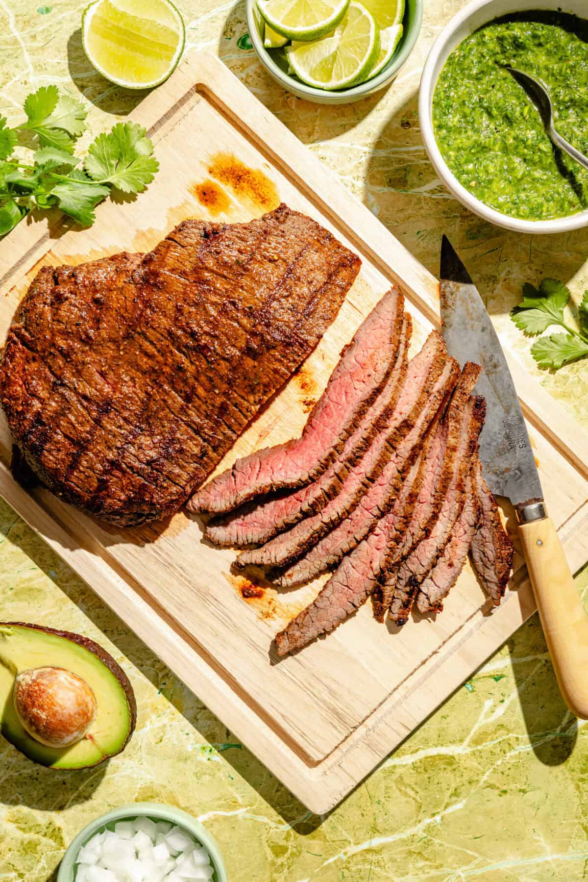 Sliced steak on wooded cutting board with knife. Other ingredients surrounding.