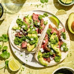 Flap Steak Tacos with Jalapeño Chimichurri assembled on plate with limes and extra sauce around.