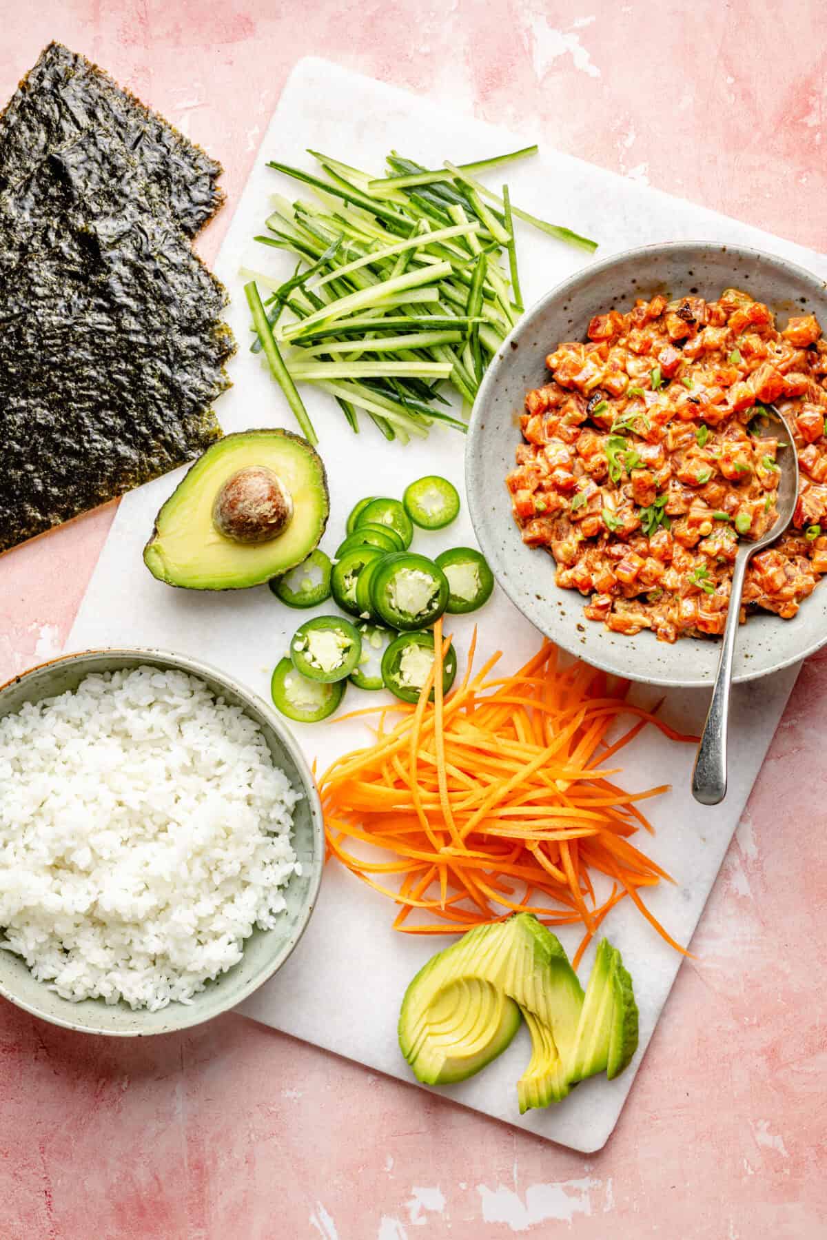 Ingredients for Spicy Tuna Hand Rolls on a white cutting board on top of a pink surface.