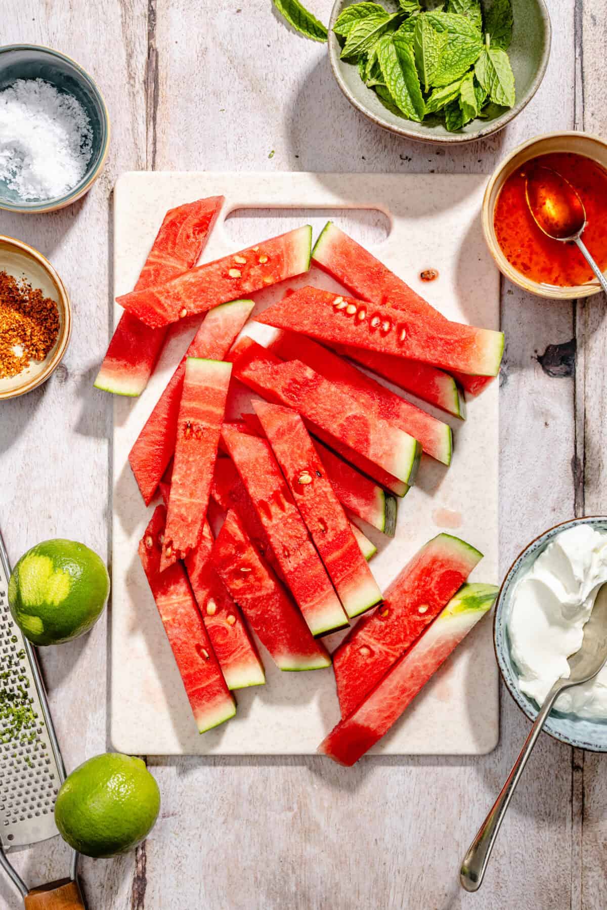 Sliced watermelon on cutting board with other ingredients scatter around.