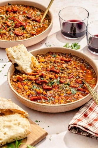 a large bowl of spanish-style lentil soup with chorizo, fresh herbs, and bread; another bowl of soup in the background